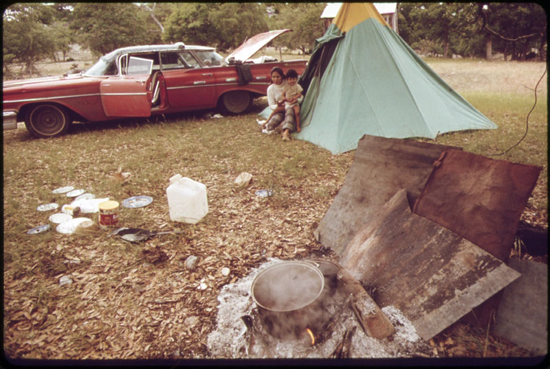 Femme et enfant d’un tondeur de moutons itinérant qui travaille pour un éleveur dans la région de Leakey et de San Antonio, au Texas. La femme cuisine ses repas sur le feu ouvert au premier plan ; la famille vit dans la tente dressée à coté de leur voiture. (Photo : Marc St. Gil, mai 1973)